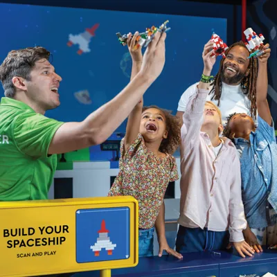 A master model builder at LEGO Discovery Center Atlanta is helping a dad and his three kids build a spaceship as part of the "Build Your Spaceship Scan and Play" attraction. The group is engaged in assembling the spaceship with colorful LEGO bricks, with the dad and kids excitedly participating in the hands-on activity. The attraction features interactive elements where participants can scan their creations for added play experiences.
