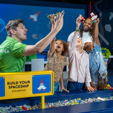 A master model builder at LEGO Discovery Center Boston is helping a dad and his three kids build a spaceship as part of the "Build Your Spaceship Scan and Play" attraction. The group is engaged in assembling the spaceship with colorful LEGO bricks, with the dad and kids excitedly participating in the hands-on activity. The attraction features interactive elements where participants can scan their creations for added play experiences.